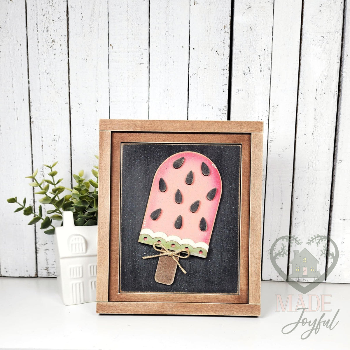 Pink watermelon popsicle 3D wood insert with black seeds, a green scalloped rind, and a twine bow on a black background, in a stained farmhouse frame.