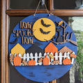 A round wooden Halloween hanging sign with a distressed blue background, featuring a yellow moon, black bats, a black cat silhouette, houses, pumpkins, leaves, and the words 'Home Spooky Home' in raised wood letters. The sign is displayed hanging on a brown wooden door.