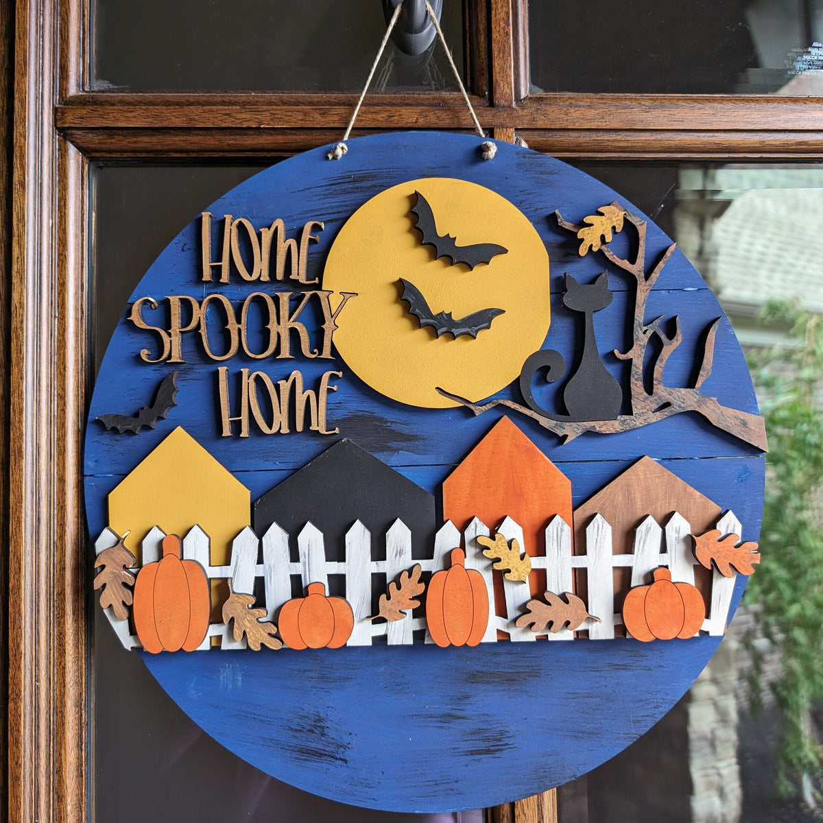 A round wooden Halloween hanging sign with a distressed blue background, featuring a yellow moon, black bats, a black cat silhouette, houses, pumpkins, leaves, and the words 'Home Spooky Home' in raised wood letters. The sign is displayed hanging on a brown wooden door.