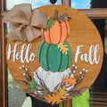 Round wooden 'Hello Fall' hanging sign with stacked pumpkins, burlap bow, and autumn leaves, displayed on a front door.