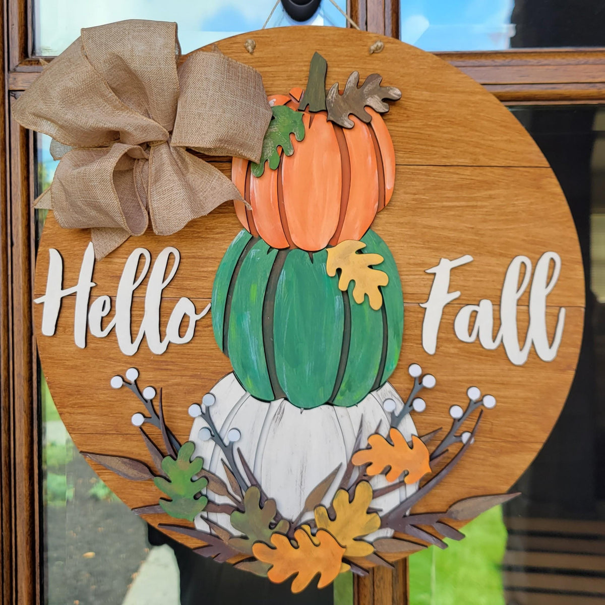 Round wooden 'Hello Fall' hanging sign with stacked pumpkins, burlap bow, and autumn leaves, displayed on a front door.