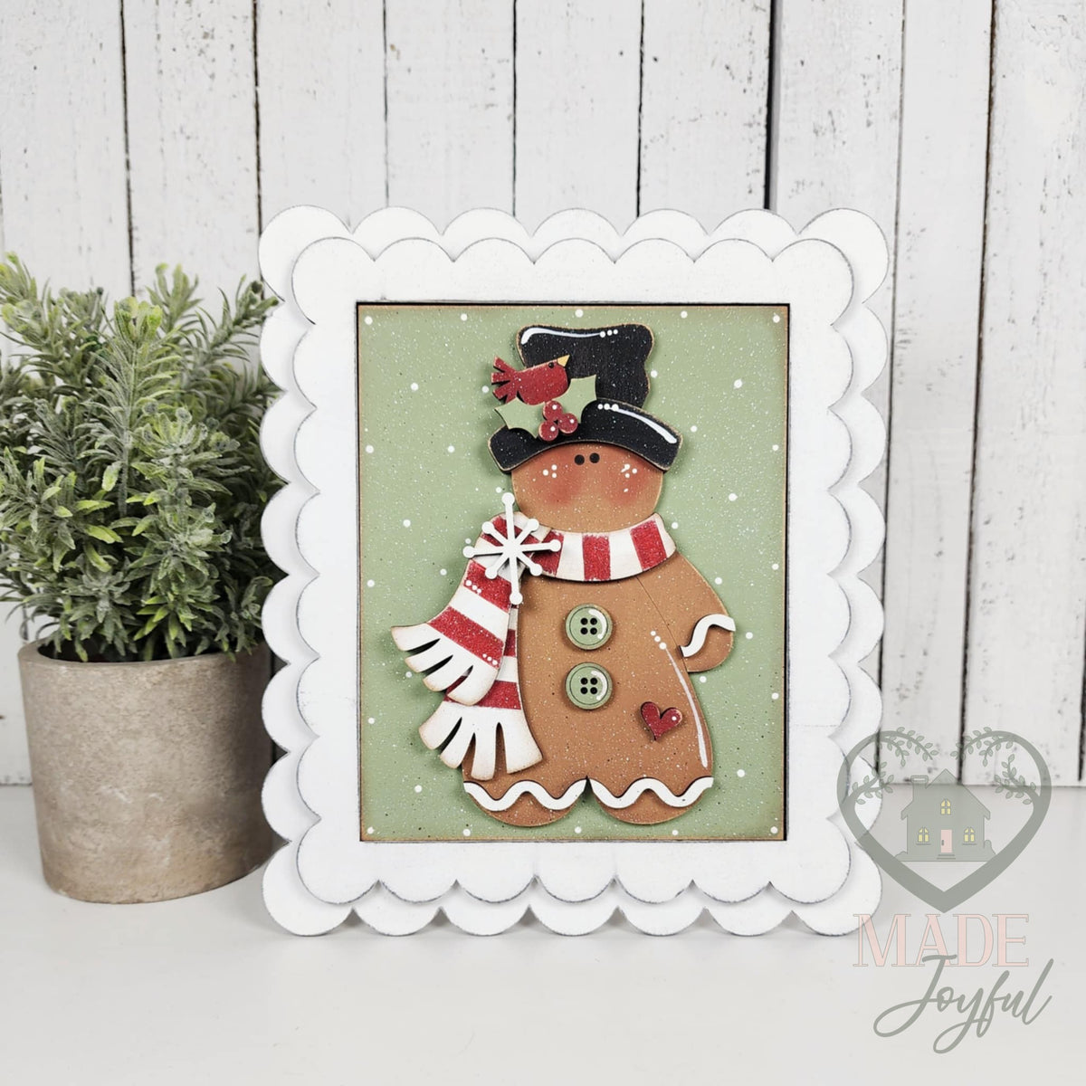 Gingerbread friend in a black top hat with a cardinal and red-striped scarf on a sage green polka-dot background, shown in a white scalloped frame.