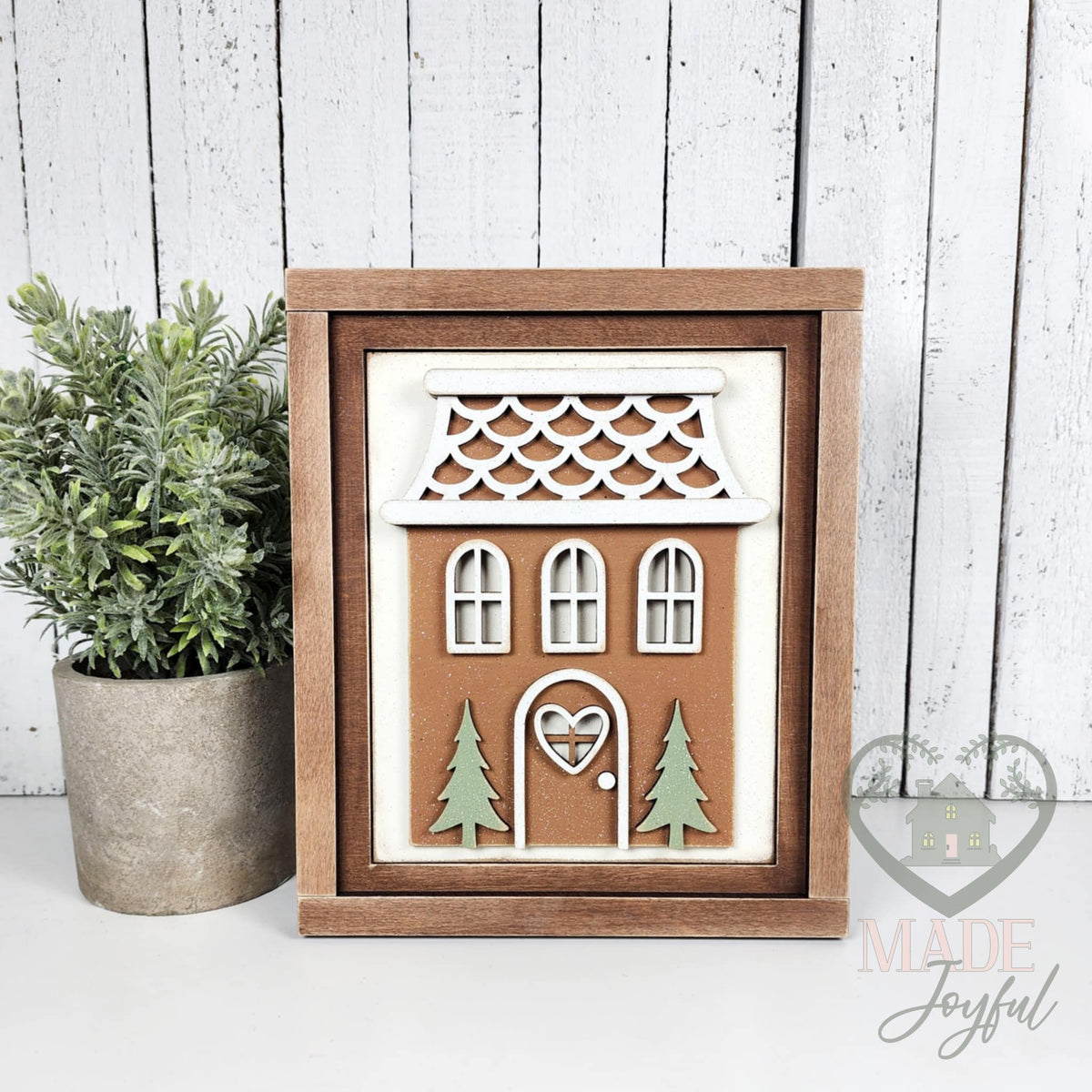 Brown gingerbread house 3D wood insert with white scalloped icing roof, arched windows, and green trees in a stained 7x8 farmhouse frame, hand-painted in Brentwood, TN.