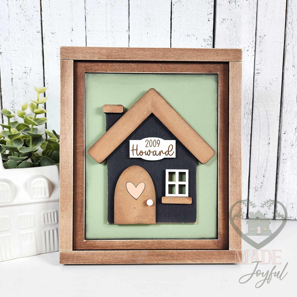 Black 3D wood house insert with a wood roof, arched door with heart, and a 2009 Howard family name plaque on a sage green background in a stained farmhouse frame.