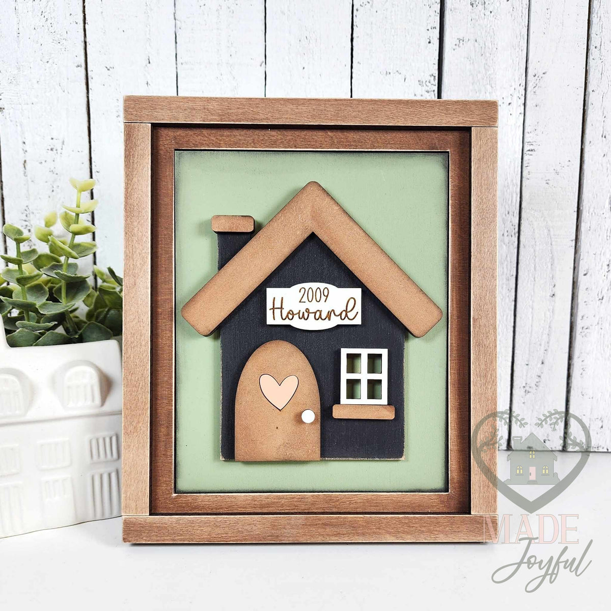 Black 3D wood house insert with a wood roof, arched door with heart, and a 2009 Howard family name plaque on a sage green background in a stained farmhouse frame.