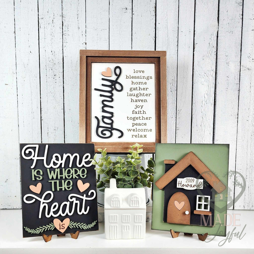 Home Is Where the Heart Is insert trio for interchangeable farmhouse frames: a Family word-list sign in a stained wood frame with a black home-heart script sign and a black house with a family name plaque on stands.