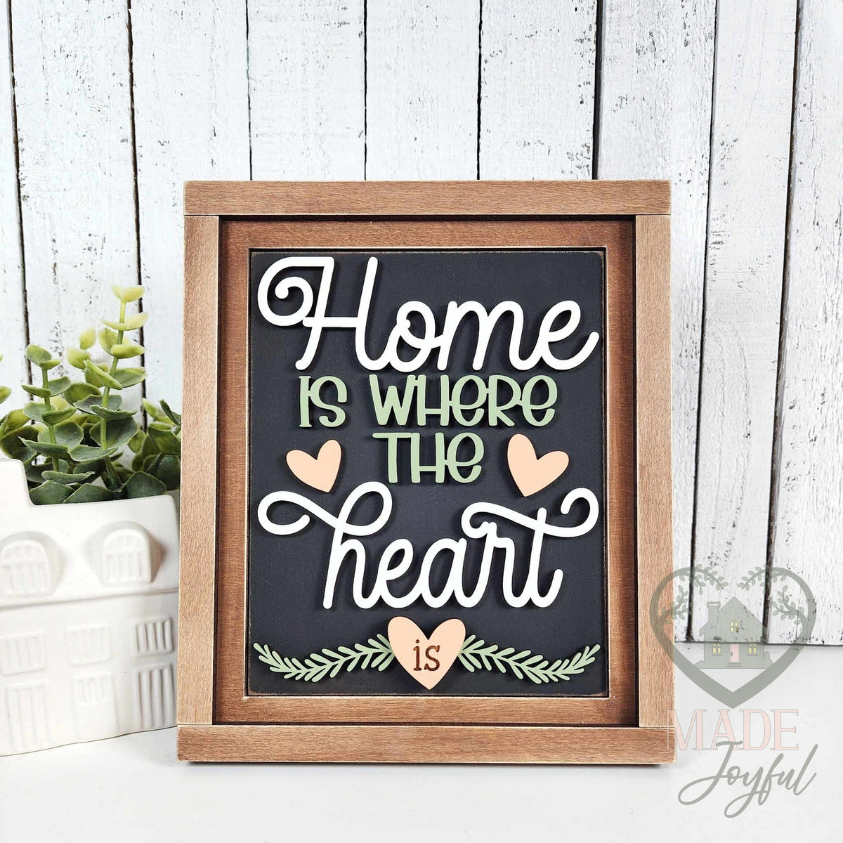 Home Is Where the Heart Is 3D wood script insert in white and sage with blush hearts and greenery on a black background, in a stained farmhouse frame.