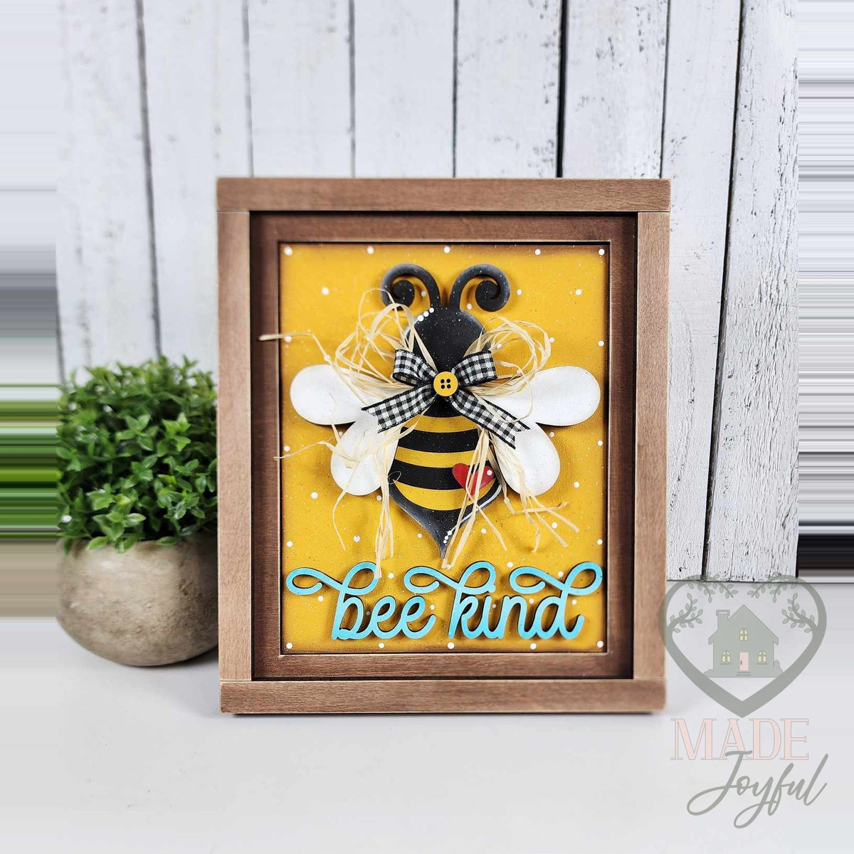 Bee Kind 3D wood insert with a large striped bee, raffia, buffalo-check bow, and teal script on a yellow polka-dot background in a stained farmhouse frame.