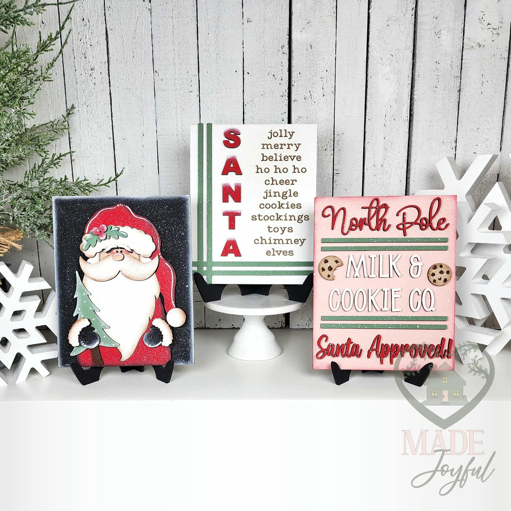 North Pole Santa insert trio for interchangeable farmhouse frames: a SANTA word-list sign on a cake stand with a woodland Santa and a North Pole Milk & Cookie Co. sign on stands, hand-painted in Brentwood, TN.