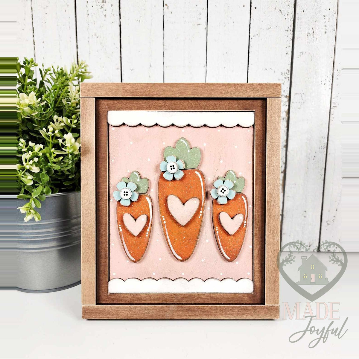Three orange carrots with heart centers and flower-button tops on a pink scalloped background, 3D wood insert in a stained farmhouse frame.