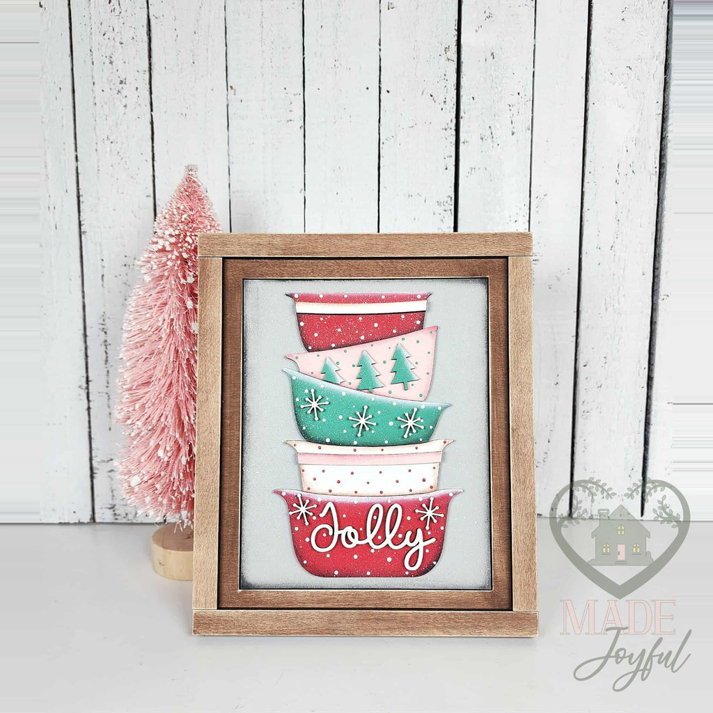 Stack of retro mixing bowls in red, mint, and pink with trees and Jolly script, 3D wood insert in a stained farmhouse frame beside a pink bottle-brush tree.
