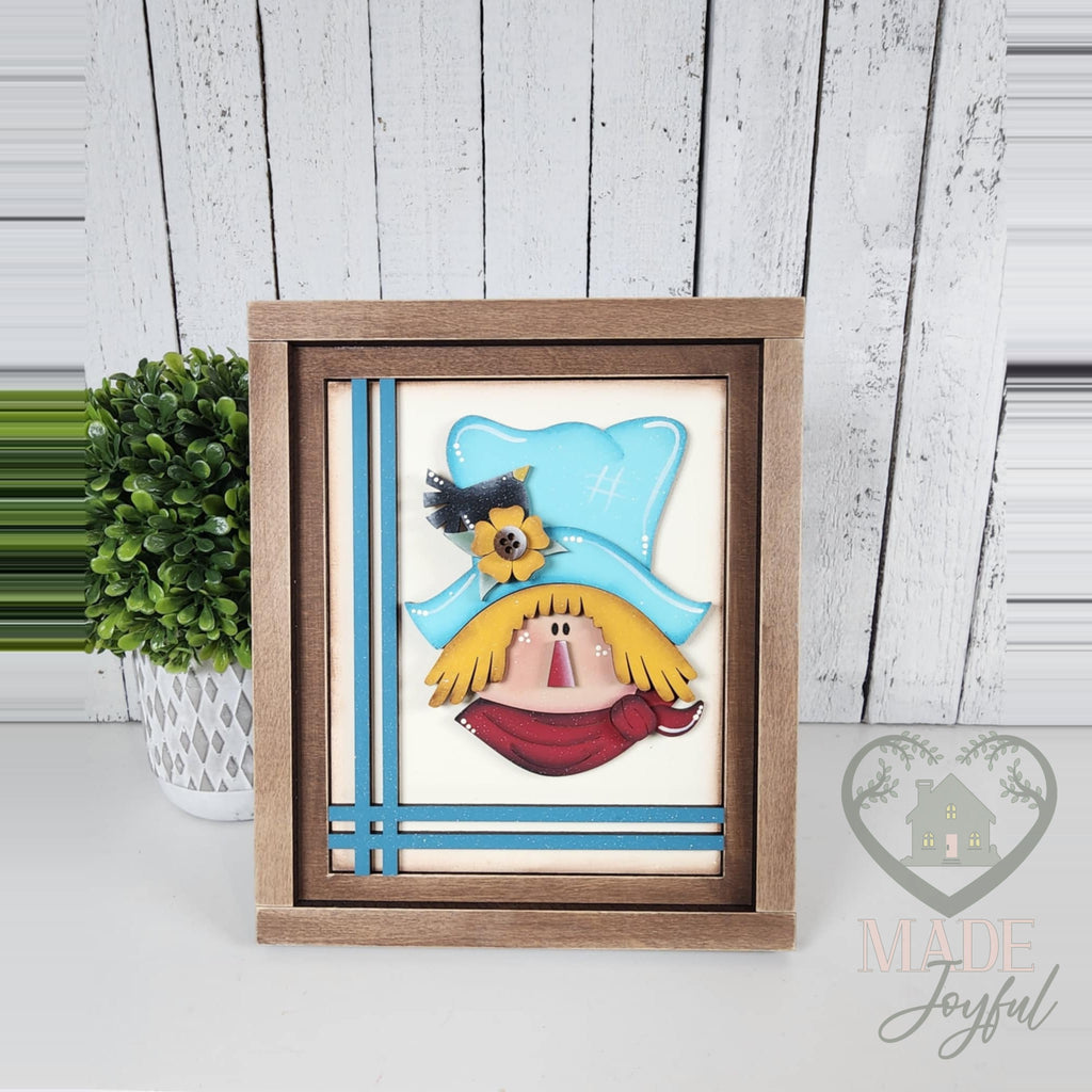 Scarecrow face 3D wood insert with a teal hat, sunflower, crow, yellow straw hair, and red scarf with teal stripes, in a stained farmhouse frame.