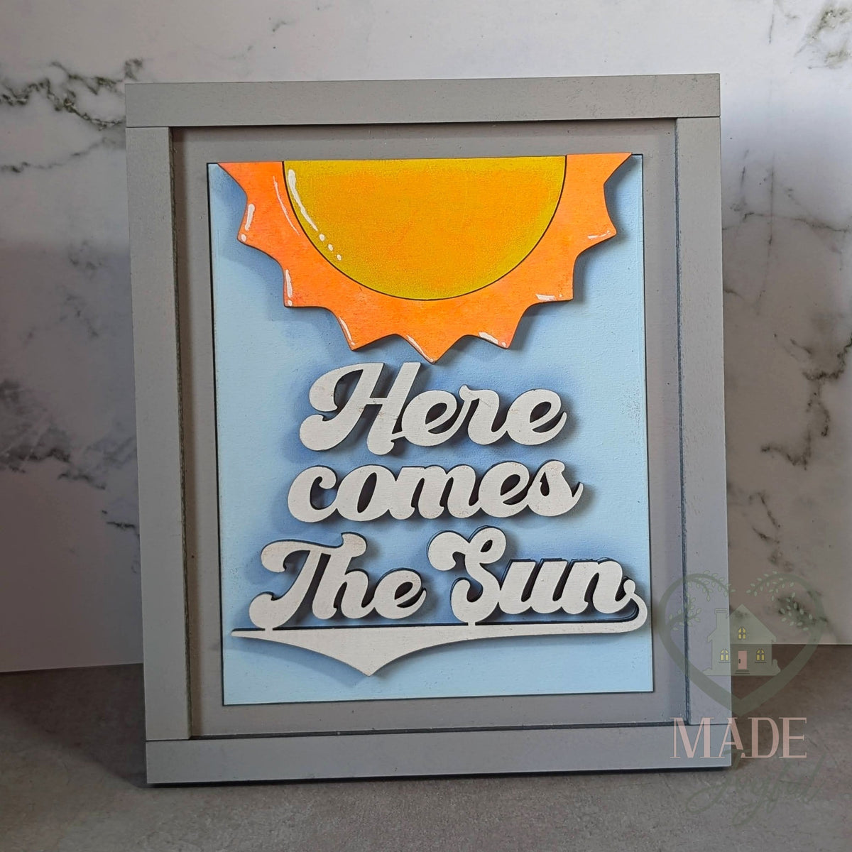 Here Comes the Sun 3D wood insert with a glossy orange sun and white script on a sky blue background, in a gray interchangeable farmhouse frame.