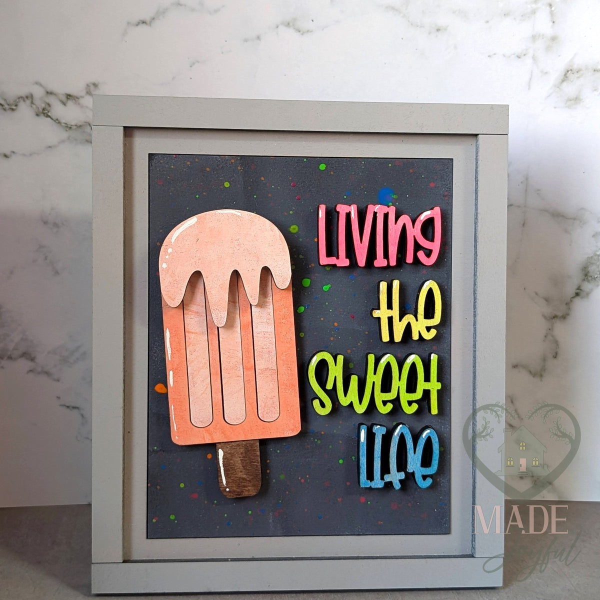 Living the Sweet Life 3D wood insert with a peach dripping popsicle and bright pink, yellow, green, and blue lettering on a black splatter background, in a gray farmhouse frame.