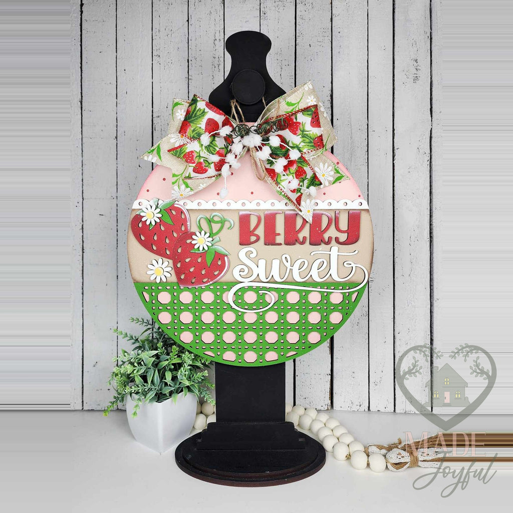 Berry Sweet strawberry door hanger displayed on a black stand beside greenery on a white shiplap shelf.