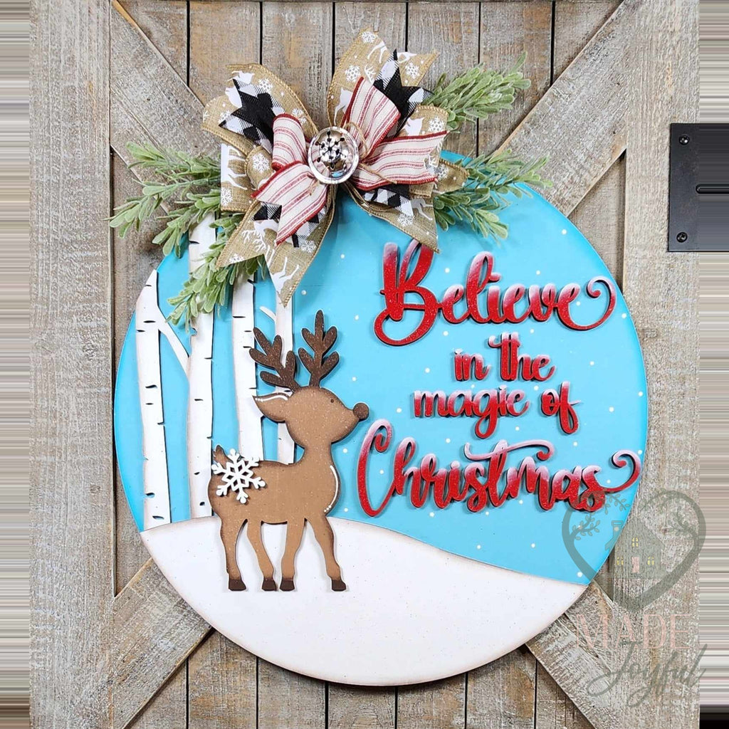 Believe in the Magic of Christmas round door hanger with a 3D wood reindeer, white birch trees, and red script on a blue sky, topped with a plaid bow and greenery, hand-painted in Brentwood, TN.