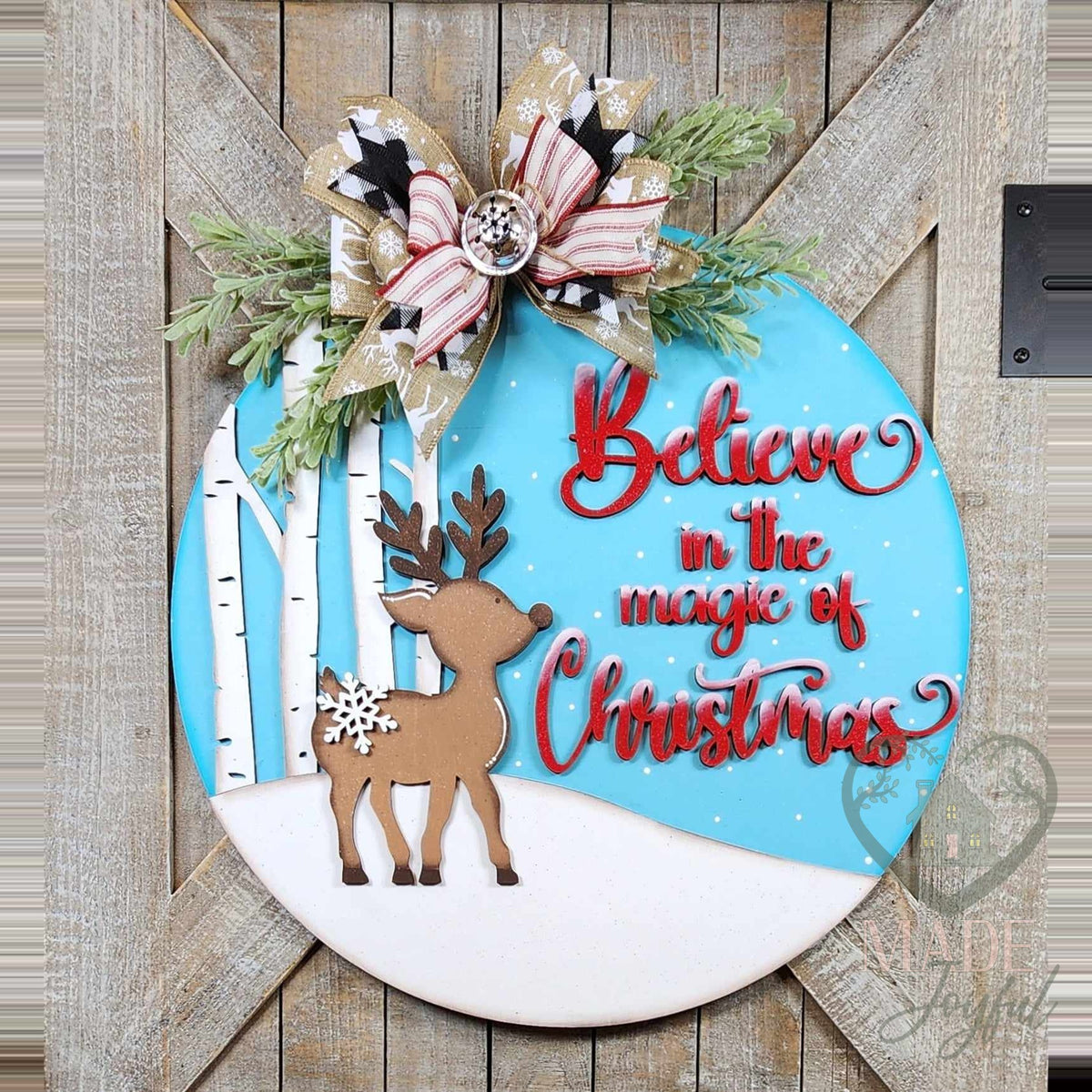 Believe in the Magic of Christmas round door hanger with a 3D wood reindeer, white birch trees, and red script on a blue sky, topped with a plaid bow and greenery, hand-painted in Brentwood, TN.