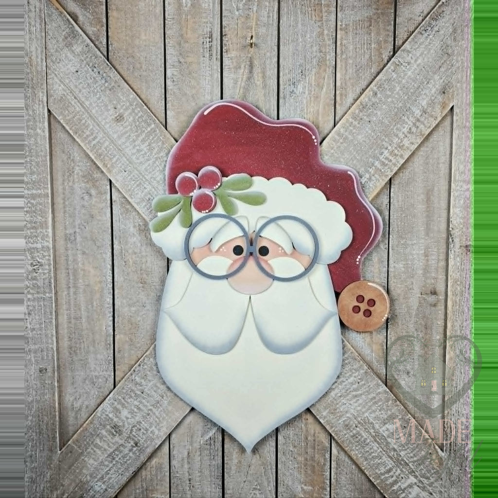 Santa face 3D wood door hanger with round glasses, holly, a red hat with a button, and a layered white beard, hand-painted in Brentwood, TN, shown on a barn door.