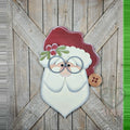 Santa face 3D wood door hanger with round glasses, holly, a red hat with a button, and a layered white beard, hand-painted in Brentwood, TN, shown on a barn door.