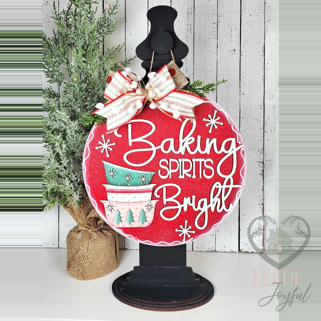 Baking Spirits Bright door hanger displayed on a black stand beside a small tree on a white shelf.