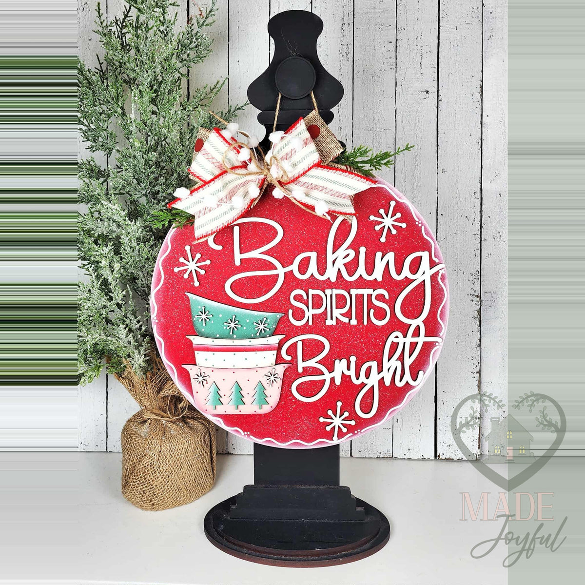 Baking Spirits Bright door hanger displayed on a black stand beside a small tree on a white shelf.