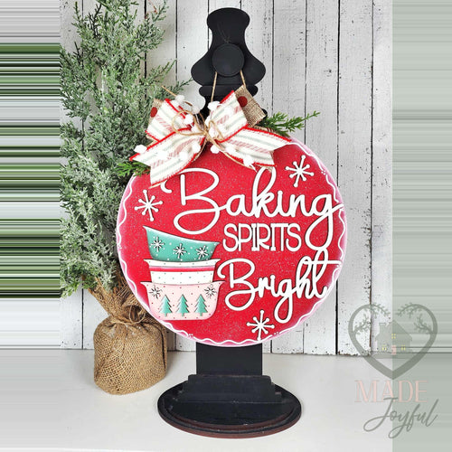 Baking Spirits Bright door hanger displayed on a black stand beside a small tree on a white shelf.