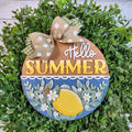 Hello Summer round wood door hanger with yellow lettering, a 3D lemon, green leaves, and white blossoms on blue, framed by greenery, hand-painted in Brentwood, TN.