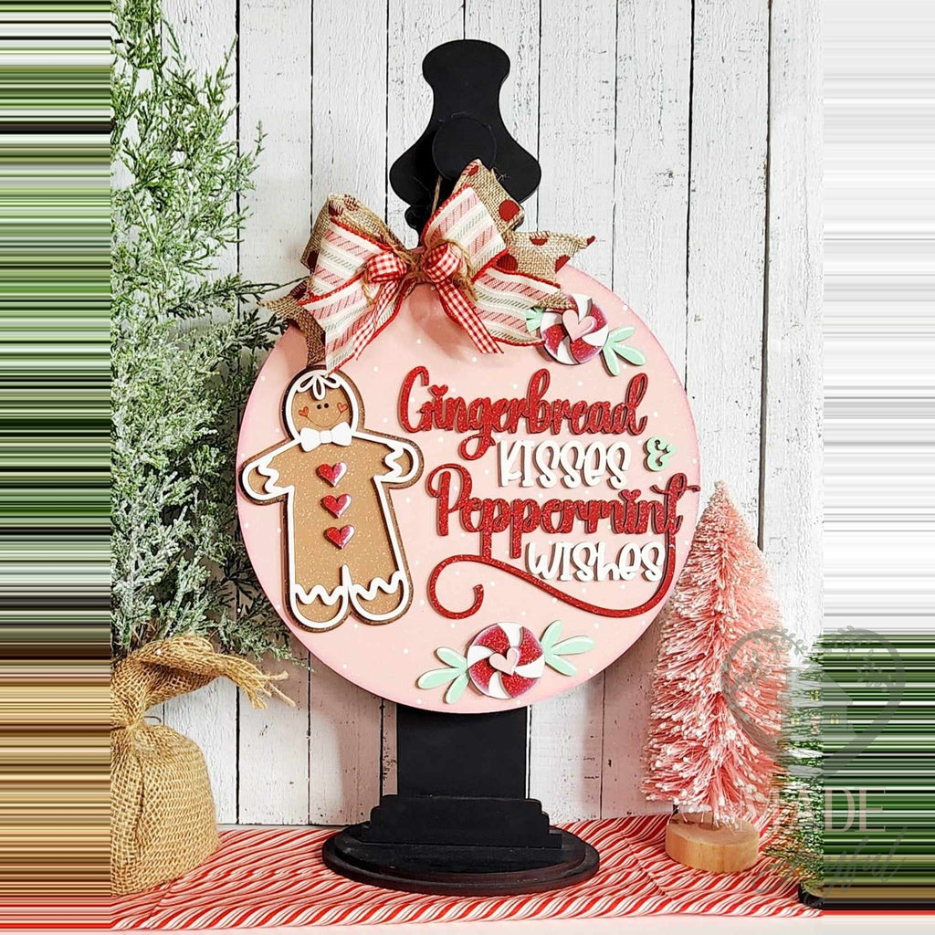 Gingerbread Kisses and Peppermint Wishes door hanger on a black stand beside pink bottle-brush trees.