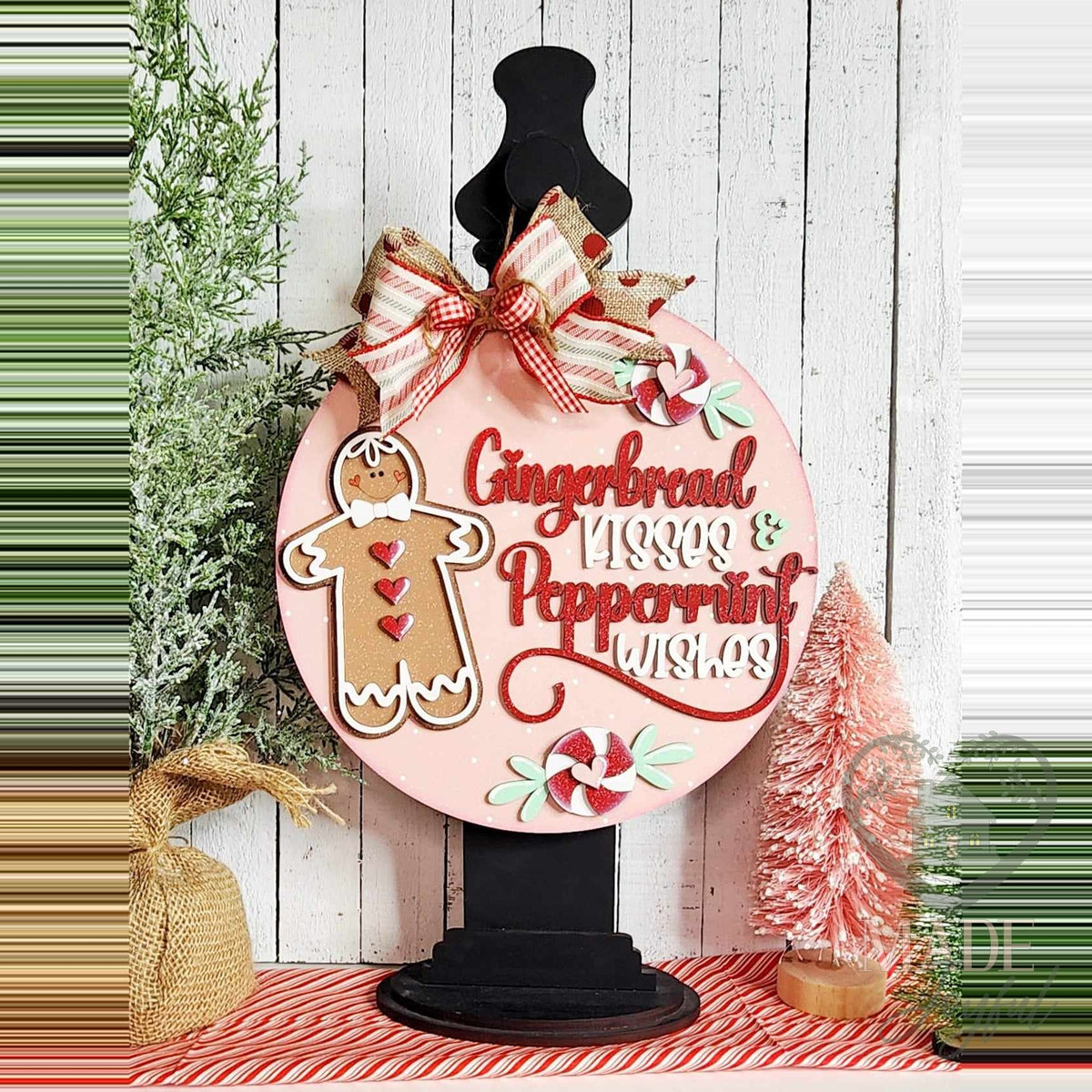 Gingerbread Kisses and Peppermint Wishes door hanger on a black stand beside pink bottle-brush trees.