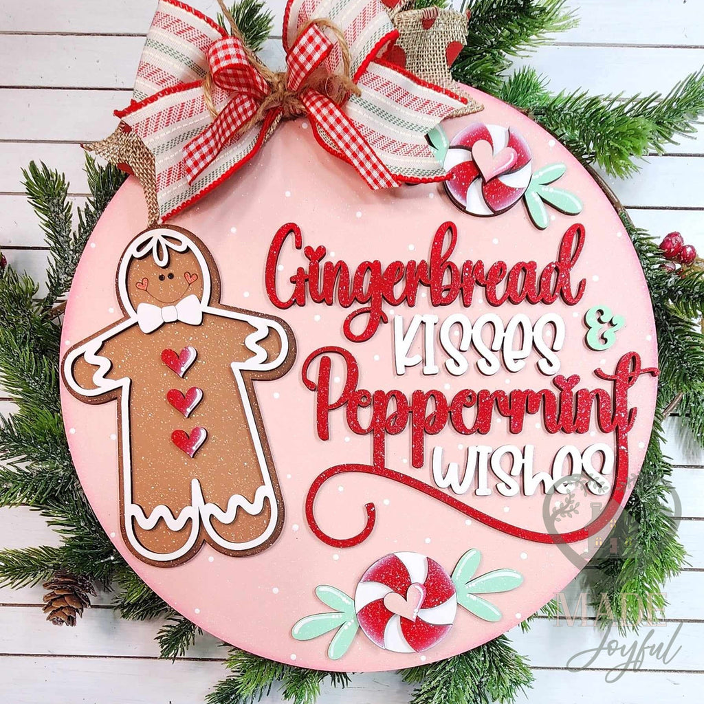 Gingerbread Kisses and Peppermint Wishes round wood door hanger with a gingerbread man, peppermints, and red script on pink, topped with a candy-stripe bow, hand-painted in Brentwood, TN.