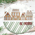 Gingerbread Village Be Merry round wood door hanger with brown gingerbread houses, white icing roofs, and a sage striped band with red Be Merry lettering, hand-painted in Brentwood, TN.