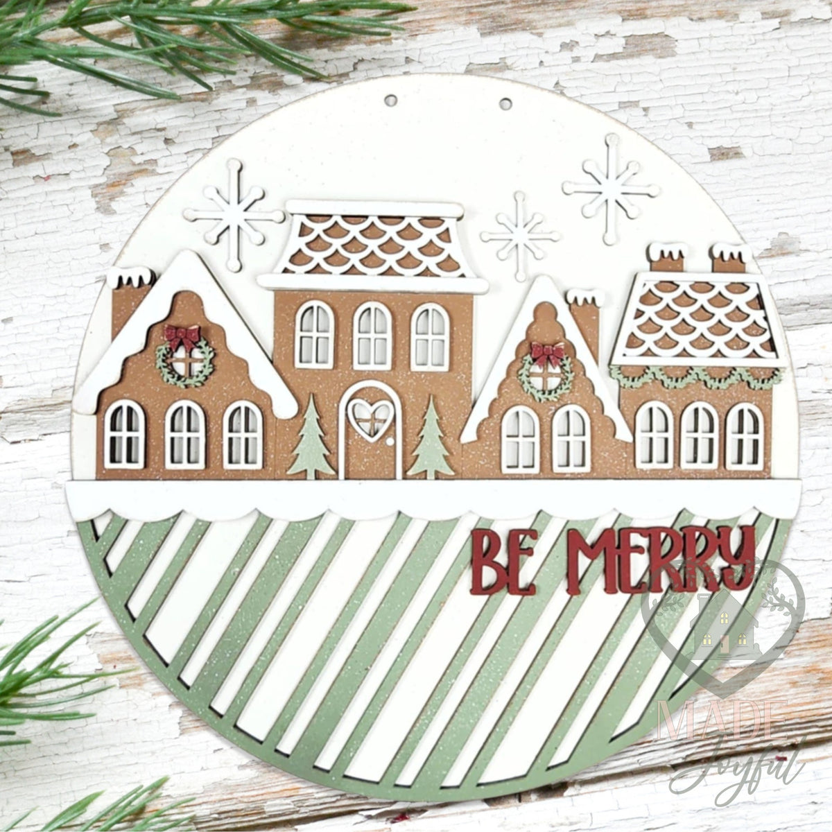 Gingerbread Village Be Merry round wood door hanger with brown gingerbread houses, white icing roofs, and a sage striped band with red Be Merry lettering, hand-painted in Brentwood, TN.