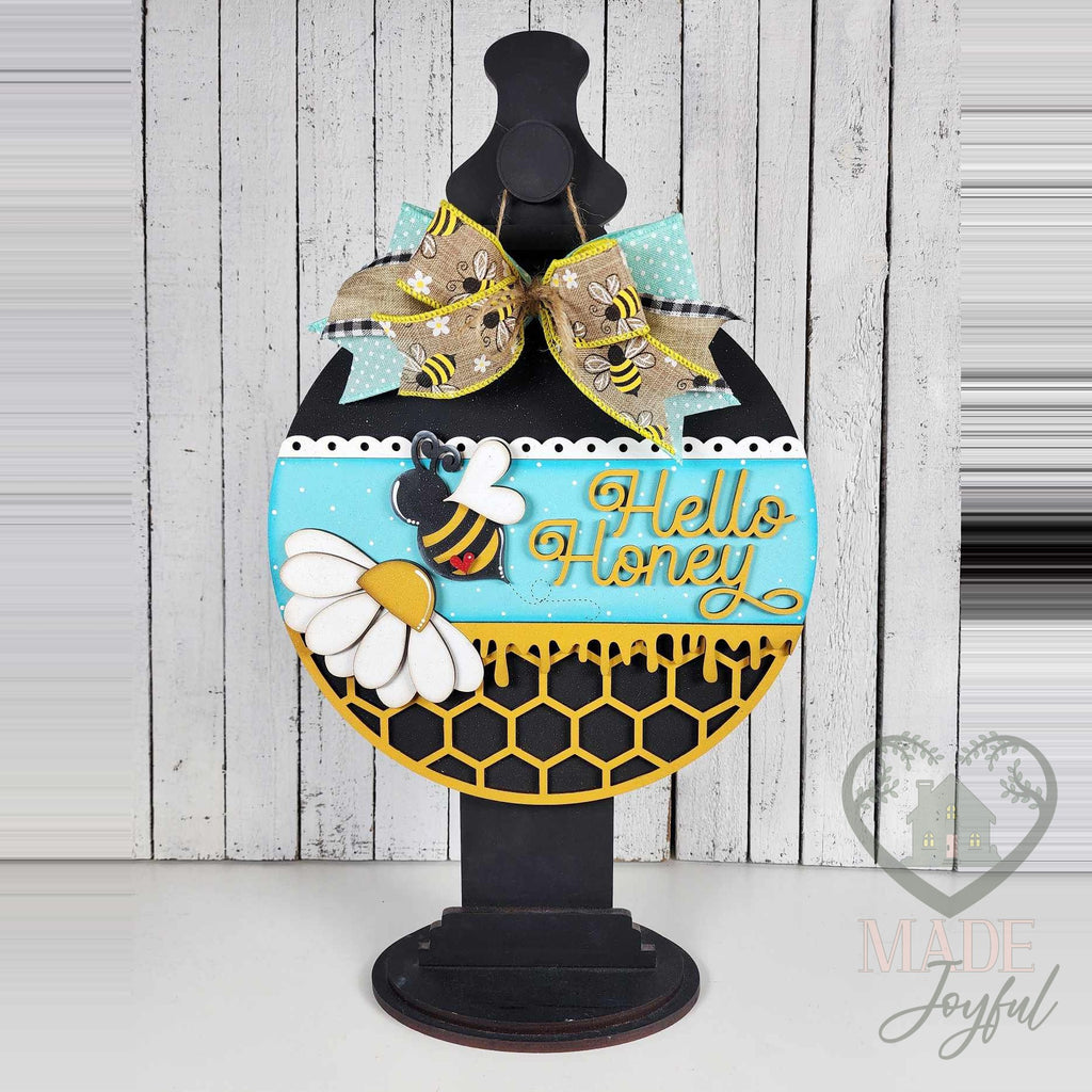 Hello Honey bee door hanger displayed on a black stand on a white shelf.