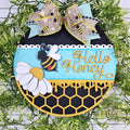 Hello Honey round wood door hanger with a bumble bee, white daisy, turquoise band, and black honeycomb with dripping honey, topped with a bee print bow, hand-painted in Brentwood, TN.