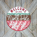 Welcome to the North Pole round wood door hanger with red lettering, a green tree band, white script, and candy-stripe snowflakes, on a barn door, hand-painted in Brentwood, TN.