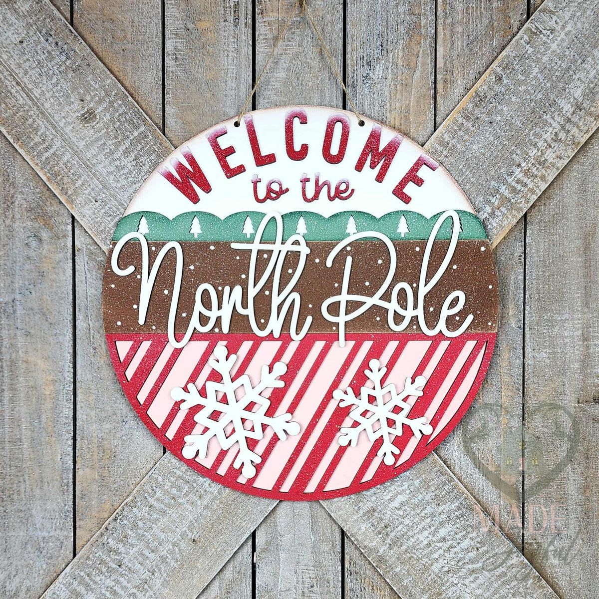 Welcome to the North Pole round wood door hanger with red lettering, a green tree band, white script, and candy-stripe snowflakes, on a barn door, hand-painted in Brentwood, TN.
