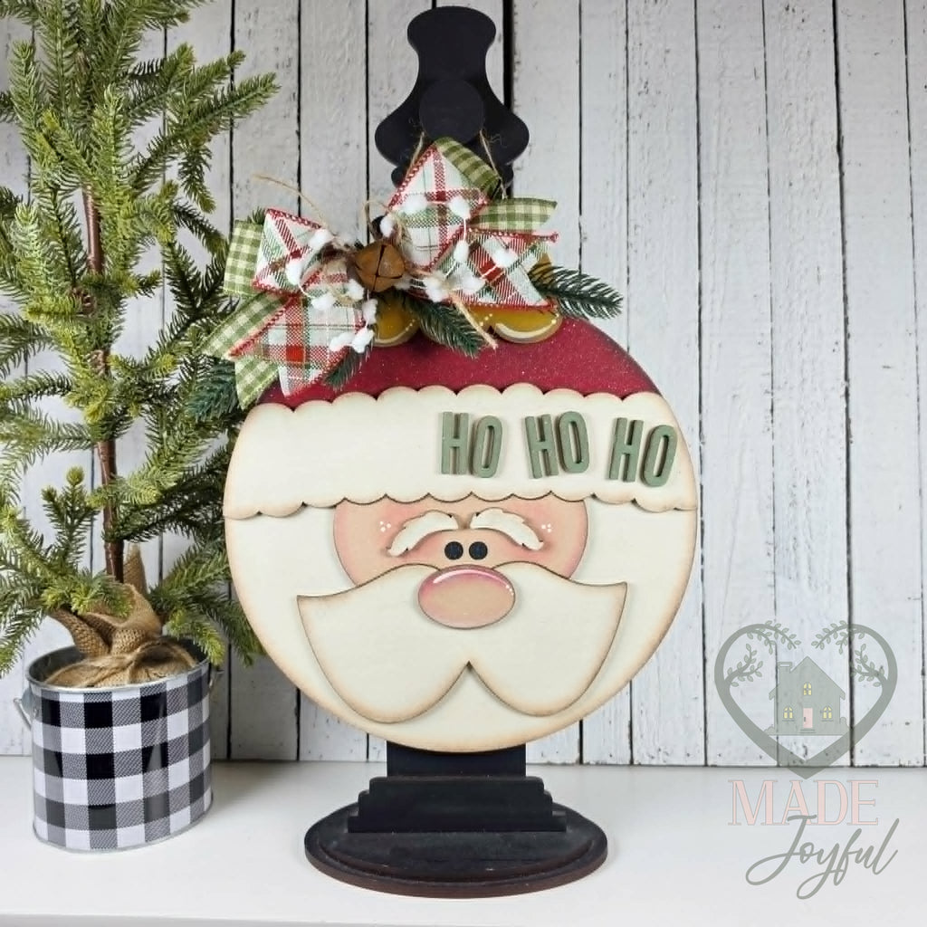 Ho Ho Ho Santa ornament-shaped wood door hanger with a red hat, rosy cheeks, and white beard, displayed on a black stand with a plaid bow, hand-painted in Brentwood, TN.