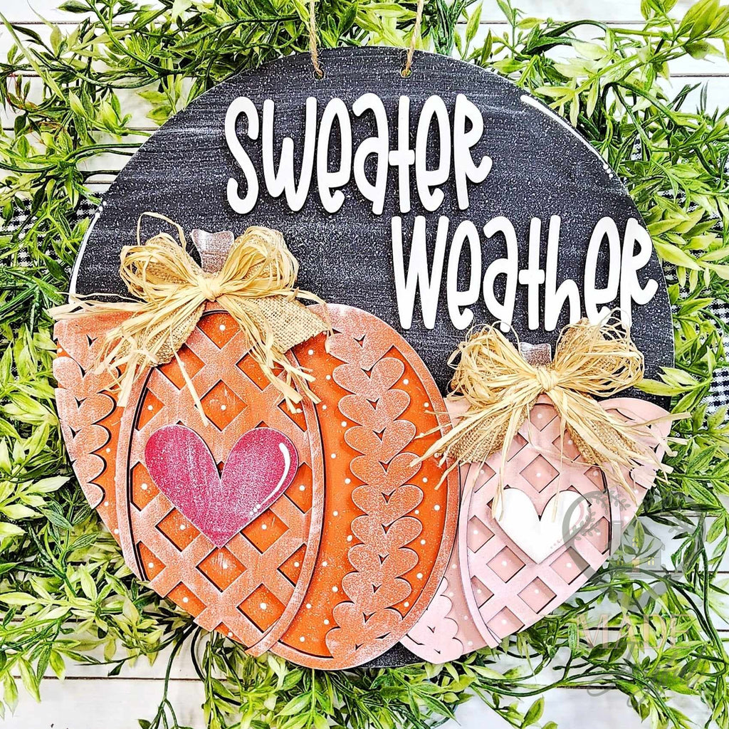 Sweater Weather round wood door hanger with knit-pattern orange and blush pumpkins, hearts, and raffia bows under white lettering on black, hand-painted in Brentwood, TN.