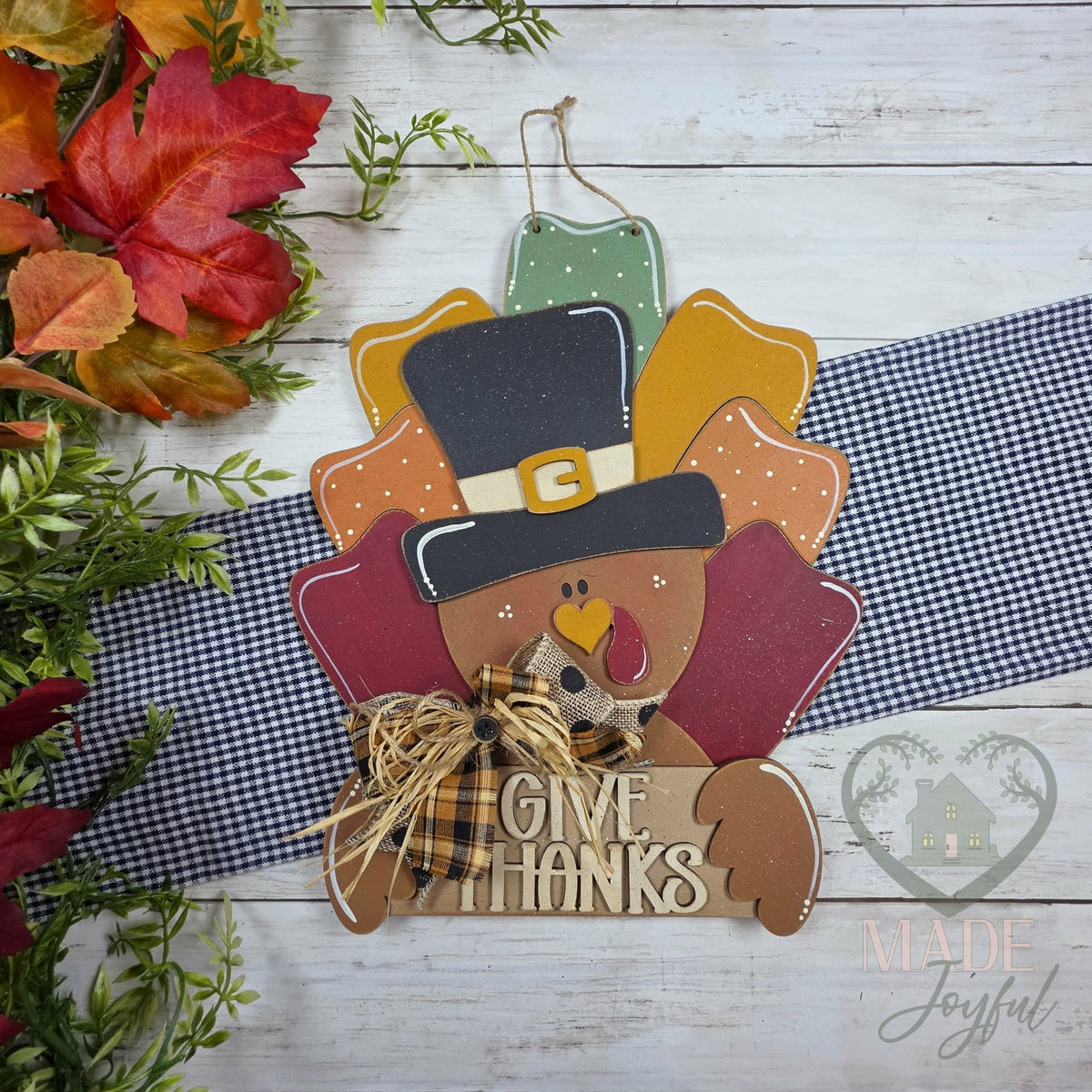 Give Thanks pilgrim turkey 3D wood door hanger with a buckled hat, plaid bow, and layered fall feathers, styled with autumn leaves, hand-painted in Brentwood, TN.