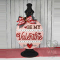 Be My Valentine round wood door hanger with red script, heart-lace borders, and a heart and arrow on pink, displayed on a black stand with a gingham bow, hand-painted in Brentwood, TN.