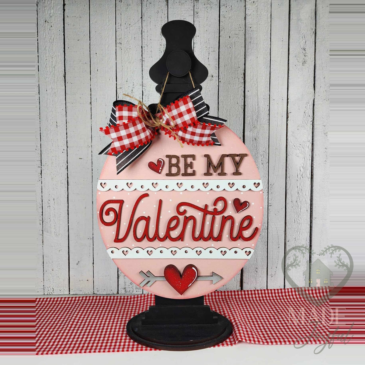 Be My Valentine round wood door hanger with red script, heart-lace borders, and a heart and arrow on pink, displayed on a black stand with a gingham bow, hand-painted in Brentwood, TN.