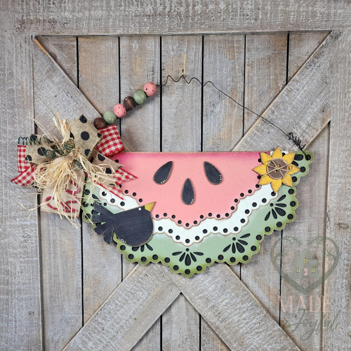 Watermelon Slice 3D wood door hanger with black seeds, a lacy green rind, a sunflower, and a crow, hung on a barn door with a beaded wire hanger, hand-painted in Brentwood, TN.