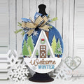 Welcome Winter Cabin wood door hanger with a white A-frame cabin, evergreen trees, and a navy sky, displayed on a black stand with a buffalo-check bow, hand-painted in Brentwood, TN.