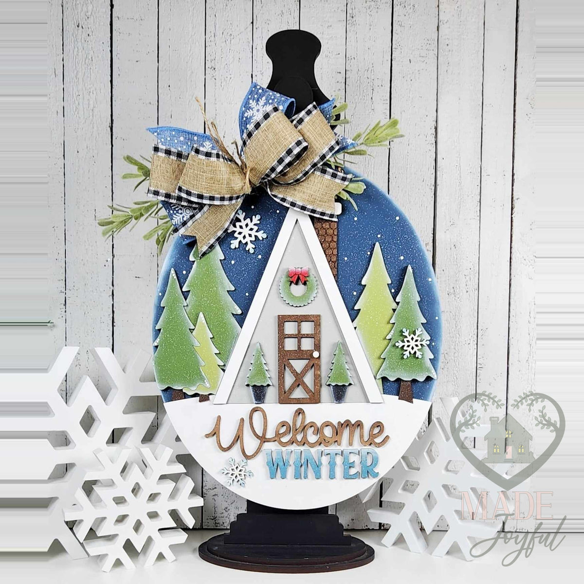 Welcome Winter Cabin wood door hanger with a white A-frame cabin, evergreen trees, and a navy sky, displayed on a black stand with a buffalo-check bow, hand-painted in Brentwood, TN.