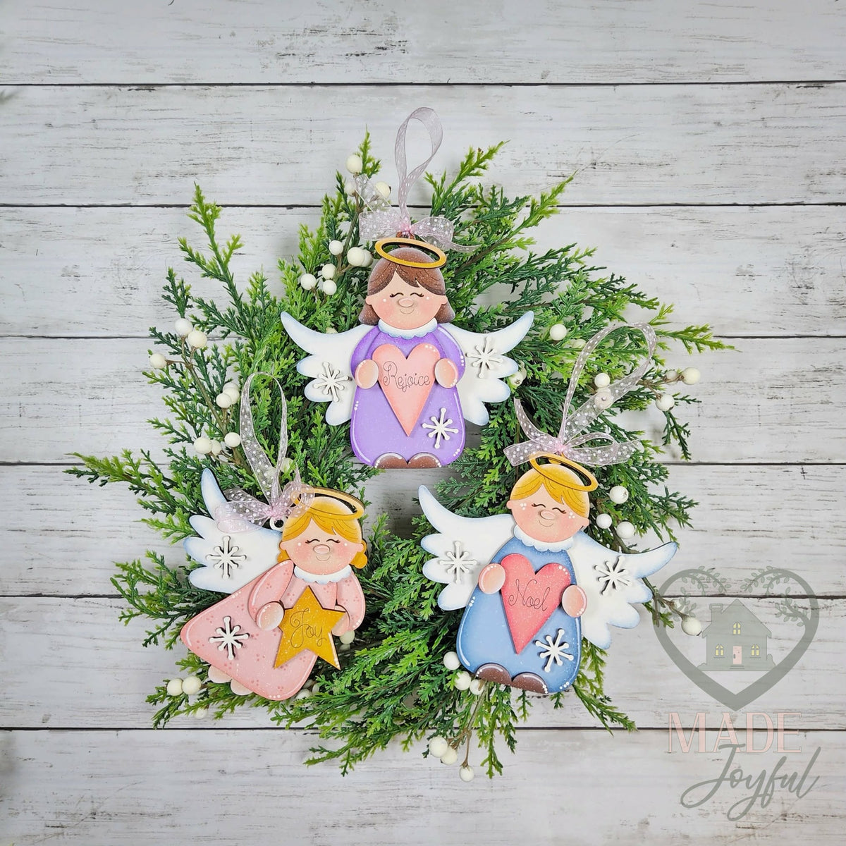 Three hand-painted 3D wood angel ornaments in purple, pink and blue with gold halos and white wings, arranged on a green wreath, hand-painted in Brentwood, TN.