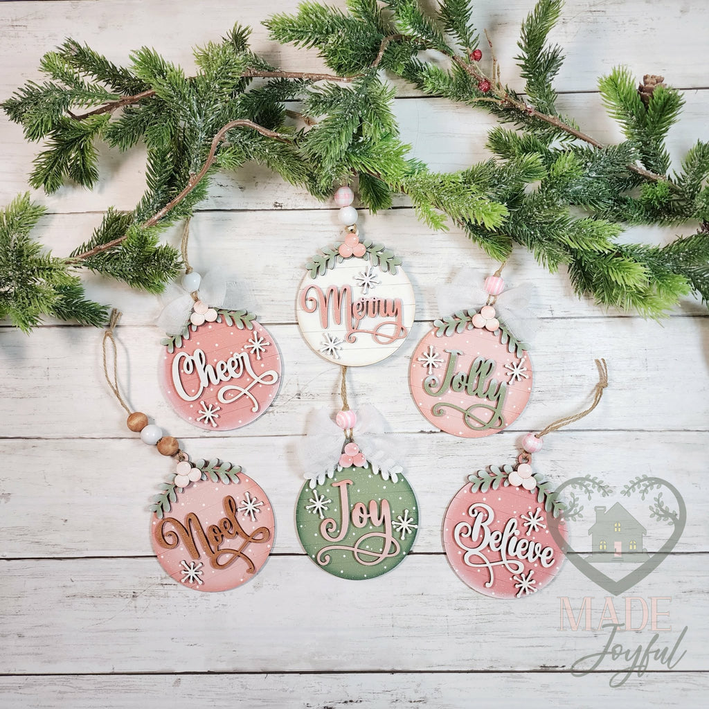 Six round wood Christmas ornaments reading Cheer, Merry, Jolly, Noel, Joy and Believe in pink, sage and cream with bead hangers, hand-painted in Brentwood, TN.