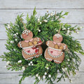 Two hand-painted gingerbread baker ornaments in pink and red aprons with trays of cookies, on a green wreath, hand-painted in Brentwood, TN.