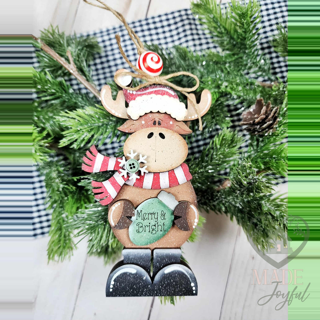 Merry and Bright moose ornament in a Santa hat, striped scarf and black boots, holding a green mitten sign, hand-painted in Brentwood, TN.