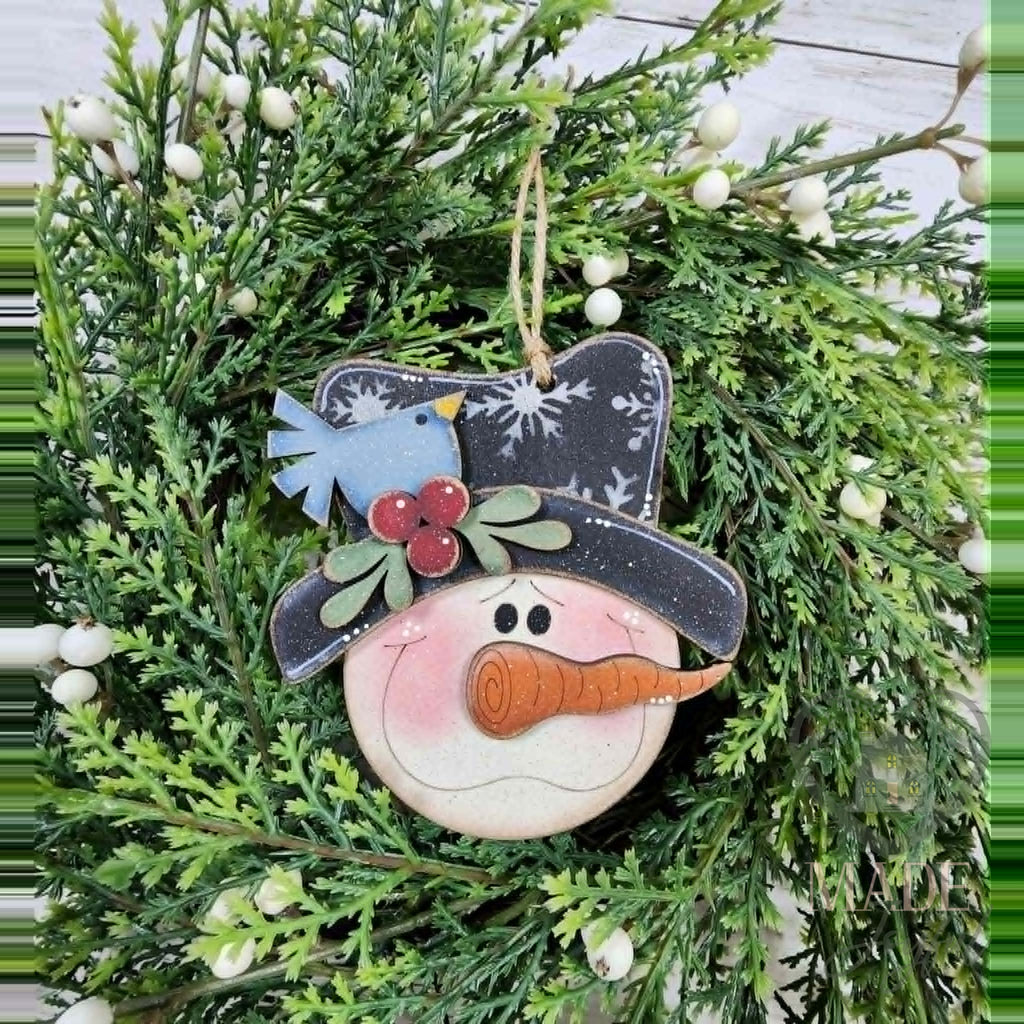 Snowman face ornament with a black snowflake top hat, bluebird, holly and carrot nose, hand-painted in Brentwood, TN.