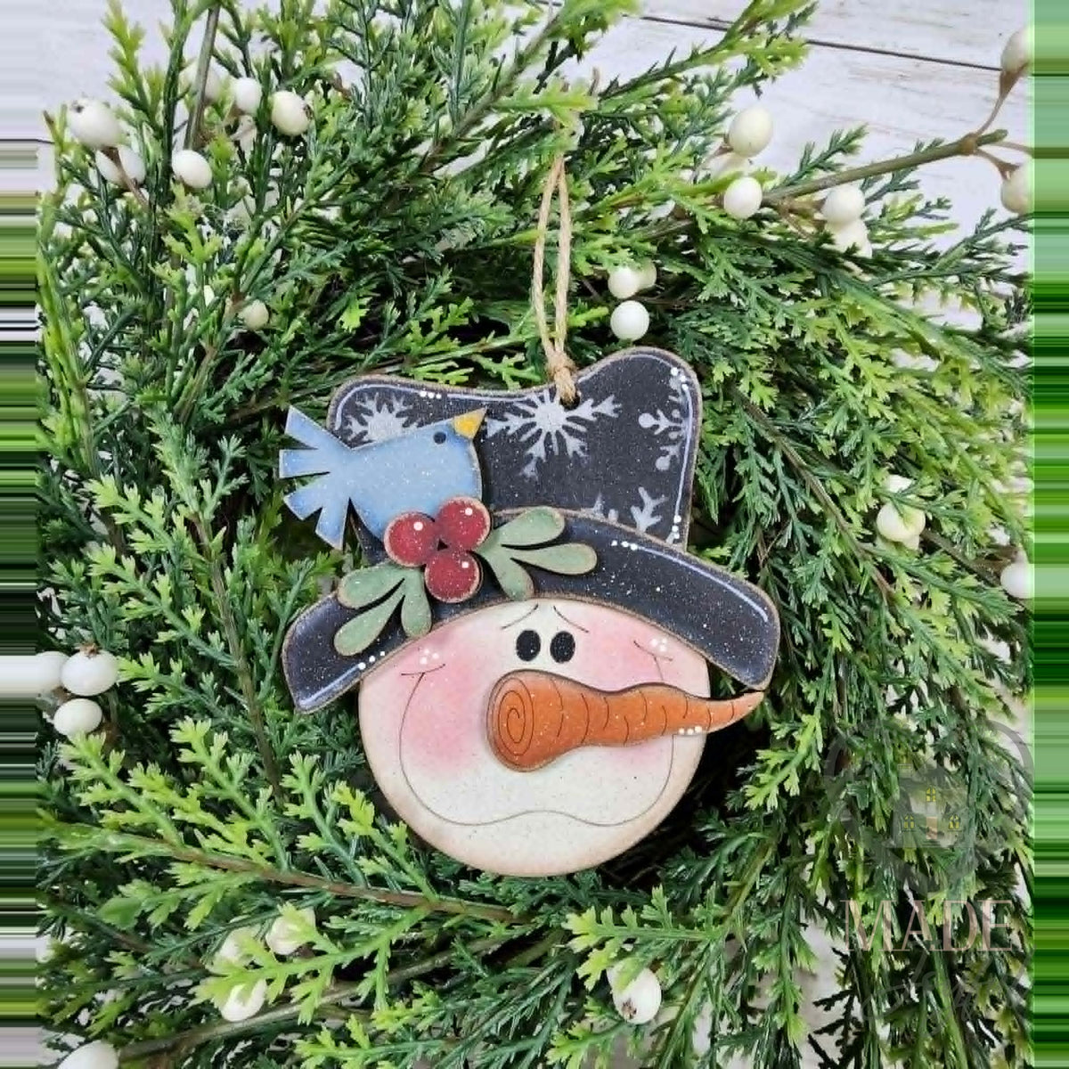 Snowman face ornament with a black snowflake top hat, bluebird, holly and carrot nose, hand-painted in Brentwood, TN.